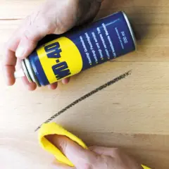 WD-40 Dose sprüht auf einen dunklen Streifen auf hellem Linoleum, der mit einem gelben Tuch abgewischt wird. So lassen sich Streifen auf Linoleum entfernen.