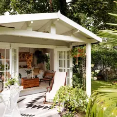 Gemütliche Schrebergarten Ideen: Ein weißes Gartenhaus mit offener Tür, Liegestuhl und Hängeampel lädt zum Verweilen ein.