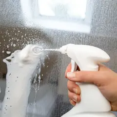 Hand sprüht Schaum auf eine beschlagene Duschwand, um sie zu reinigen. So wird blindes Bleikristall reinigen zum Kinderspiel.