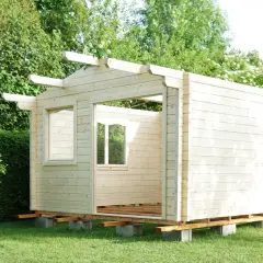 Ein Rohbau für ein Gartenhaus, das man selber bauen kann, steht im Grünen. Die Holzwände sind bereits montiert, Fensteröffnungen sind sichtbar.