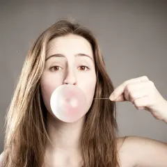 Ein Mädchen bläst einen Kaugummi auf. Sie hält einen dünnen Faden, der an der Blase befestigt ist, und denkt darüber nach, wie man Kaugummi mit Haarspray entfernen kann.