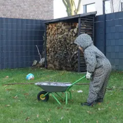 Kind schiebt Schubkarre mit Ästen am Zaun entlang. Gartengestaltung am Zaun entlang mit Holzstapel und Ball.