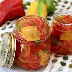 Zwei Gläser mit eingelegter Paprika, daneben frische rote, gelbe und grüne Paprika. Perfekt zum Paprika einkochen.