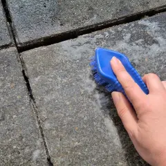 Eine Hand mit blauer Bürste reinigt Betonplatten. Essig greift den Beton an, Schaum entsteht.