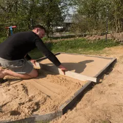 Mann richtet den sandigen Untergrund für den Sandkastenbau mit einer Wasserwaage.