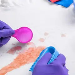 Lila Handschuhe behandeln Kirschsaftflecken auf weißem Stoff mit einer Bürste und einem Reinigungsmittel.