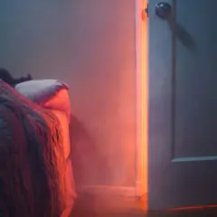 Person schläft tief unter Decken, während ein unheimliches rotes Licht durch die offene Tür fällt. Nach Brand in Wohnung schlafen ist eine Herausforderung.