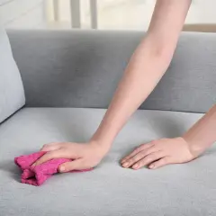 Eine Hand mit einem rosa Tuch reinigt den grauen Polsterstoff, um einen Fettfleck aus Polster zu entfernen.