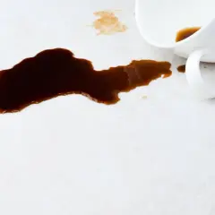 Ein umgekippter Becher hat dunklen Kaffee auf einen hellen Teppich verschüttet. So kann man Kaffee aus Teppich entfernen.