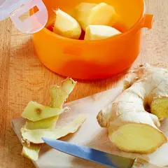 Frisch geschältes Ingwerstück liegt neben einem Messer auf einem Holzbrett. Ein orangefarbener Behälter mit weiteren Ingwerstücken ist bereit zum **ingwer einfrieren**.