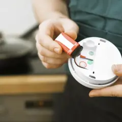 Hände tauschen eine Batterie in einem Rauchmelder aus. Sicherheit geht vor, darum den Rauchmelder regelmäßig warten.