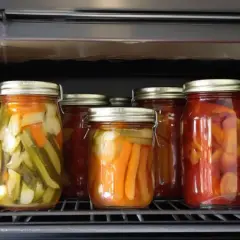 Sechs Gläser mit eingelegtem Gemüse, darunter Gurken und Paprika, stehen auf einem Rost im Backofen. Das ist eine Methode zum **einwecken im Backofen**.