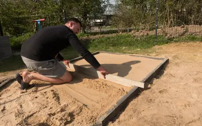 Mann richtet den sandigen Untergrund für den Sandkastenbau mit einer Wasserwaage.