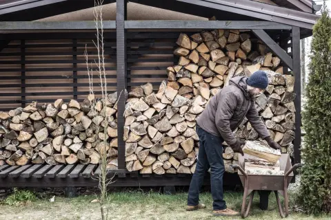 Mann lagert brennholz. Ein Mann mit Mütze und Jacke legt Holzscheite in eine Schubkarre, um sie zum Stapel zu bringen.