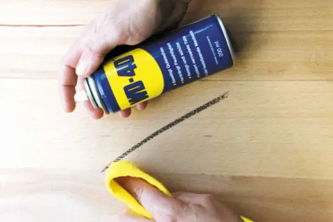 WD-40 Dose sprüht auf einen dunklen Streifen auf hellem Linoleum, der mit einem gelben Tuch abgewischt wird. So lassen sich Streifen auf Linoleum entfernen.