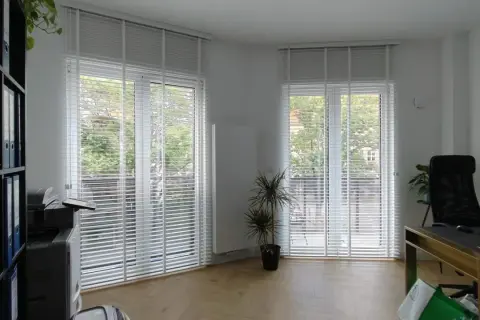 Büro mit vergilbten Plissees reinigen. Helle Räume mit viel Tageslicht, ein Schreibtisch und ein Regal.