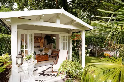 Gemütliche Schrebergarten Ideen: Ein weißes Gartenhaus mit offener Tür, Liegestuhl und Hängeampel lädt zum Verweilen ein.