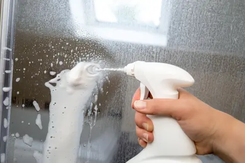 Hand sprüht Schaum auf eine beschlagene Duschwand, um sie zu reinigen. So wird blindes Bleikristall reinigen zum Kinderspiel.