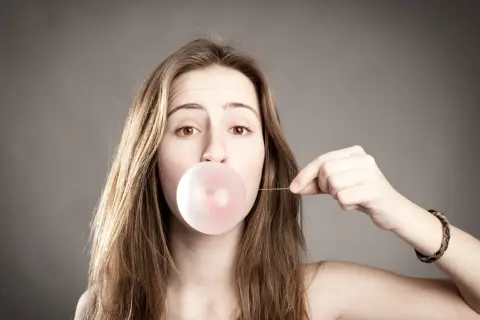 Ein Mädchen bläst einen Kaugummi auf. Sie hält einen dünnen Faden, der an der Blase befestigt ist, und denkt darüber nach, wie man Kaugummi mit Haarspray entfernen kann.