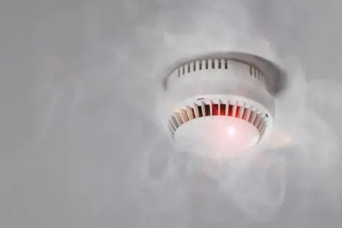 Rauchmelder mit rotem Licht und Dampf, der einen Fehlalarm durch hohe Luftfeuchtigkeit auslöst.