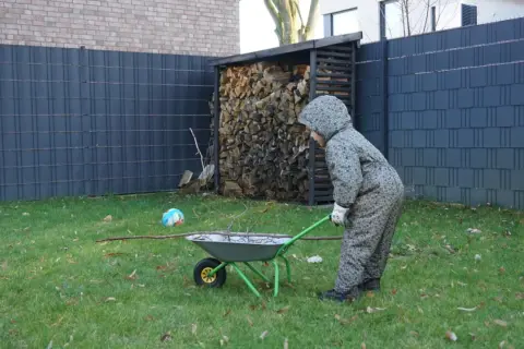 Kind schiebt Schubkarre mit Ästen am Zaun entlang. Gartengestaltung am Zaun entlang mit Holzstapel und Ball.