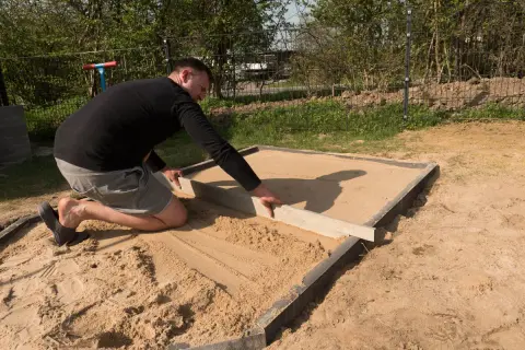Mann richtet den sandigen Untergrund für den Sandkastenbau mit einer Wasserwaage.