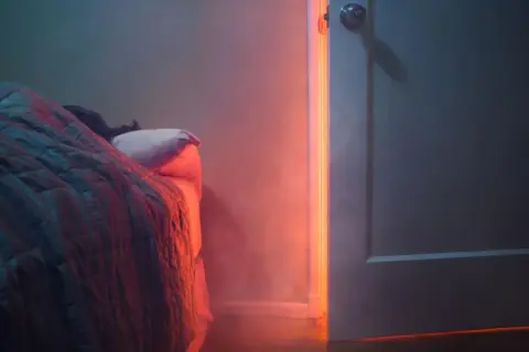 Person schläft tief unter Decken, während ein unheimliches rotes Licht durch die offene Tür fällt. Nach Brand in Wohnung schlafen ist eine Herausforderung.