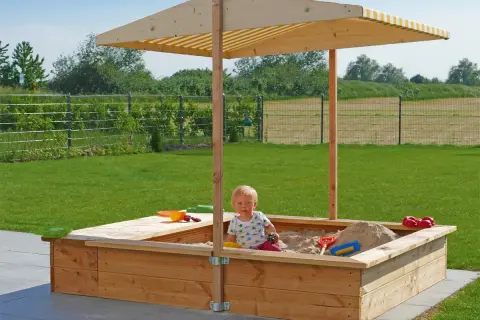 Ein Kind spielt im **Sandkasten selber bauen**. Der Holzsandkasten mit Sonnendach steht im Garten.