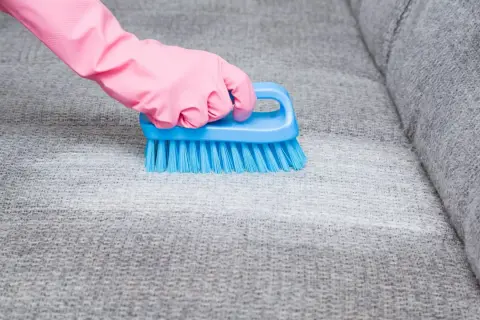Hand in pink glove uses blue brush to remove blutflecken from grey sofa fabric.