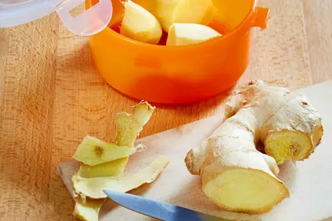Frisch geschältes Ingwerstück liegt neben einem Messer auf einem Holzbrett. Ein orangefarbener Behälter mit weiteren Ingwerstücken ist bereit zum **ingwer einfrieren**.