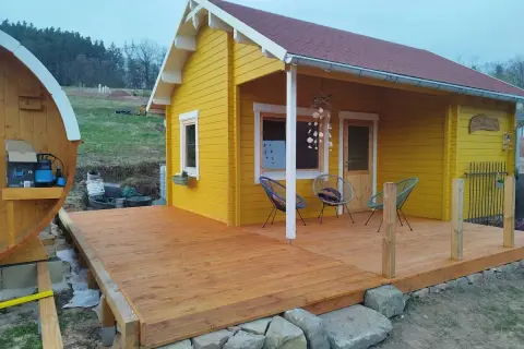 Ein gelber Holzschuppen mit Terrasse, ideal zum schuppen selber bauen. Daneben eine Holzfasssauna.