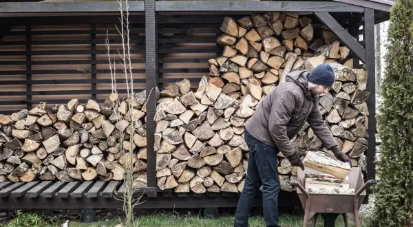 Mann lagert brennholz. Ein Mann mit Mütze und Jacke legt Holzscheite in eine Schubkarre, um sie zum Stapel zu bringen.