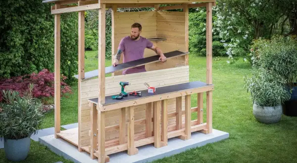 Mann baut eine Gartenbar selber. Holzkonstruktion mit Dach, Tresen und Werkzeug.