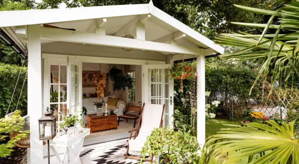 Gemütliche Schrebergarten Ideen: Ein weißes Gartenhaus mit offener Tür, Liegestuhl und Hängeampel lädt zum Verweilen ein.