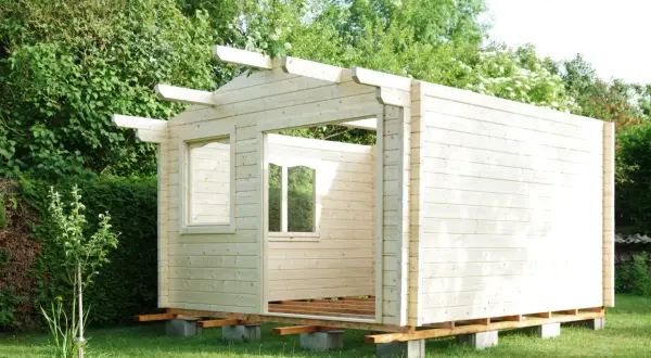 Ein Rohbau für ein Gartenhaus, das man selber bauen kann, steht im Grünen. Die Holzwände sind bereits montiert, Fensteröffnungen sind sichtbar.