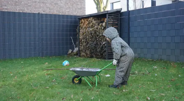 Kind schiebt Schubkarre mit Ästen am Zaun entlang. Gartengestaltung am Zaun entlang mit Holzstapel und Ball.