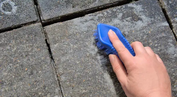 Eine Hand mit blauer Bürste reinigt Betonplatten. Essig greift den Beton an, Schaum entsteht.