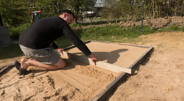 Mann richtet den sandigen Untergrund für den Sandkastenbau mit einer Wasserwaage.
