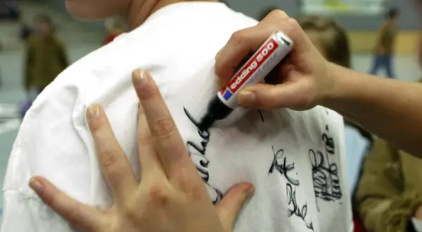 Eine Hand schreibt mit einem Edding-Filzstift Autogramme auf ein weißes T-Shirt. Tipps für hausmittel filzstift entfernen sind hier nicht zu sehen.