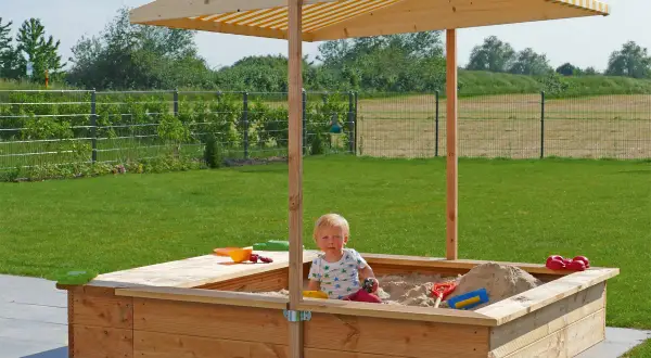 Ein Kind spielt im **Sandkasten selber bauen**. Der Holzsandkasten mit Sonnendach steht im Garten.