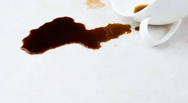 Ein umgekippter Becher hat dunklen Kaffee auf einen hellen Teppich verschüttet. So kann man Kaffee aus Teppich entfernen.