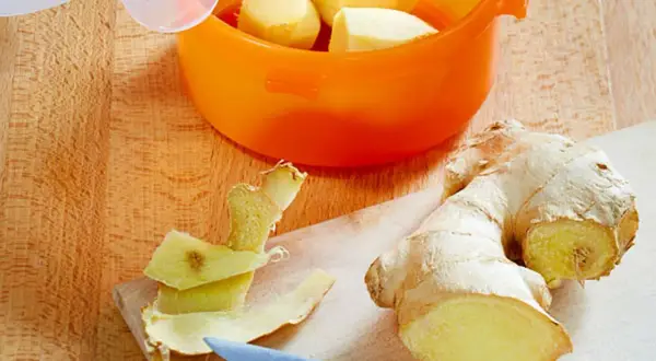 Frisch geschältes Ingwerstück liegt neben einem Messer auf einem Holzbrett. Ein orangefarbener Behälter mit weiteren Ingwerstücken ist bereit zum **ingwer einfrieren**.