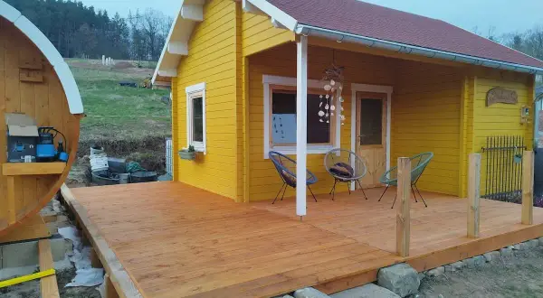Ein gelber Holzschuppen mit Terrasse, ideal zum schuppen selber bauen. Daneben eine Holzfasssauna.