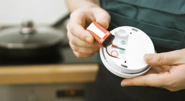 Hände tauschen eine Batterie in einem Rauchmelder aus. Sicherheit geht vor, darum den Rauchmelder regelmäßig warten.