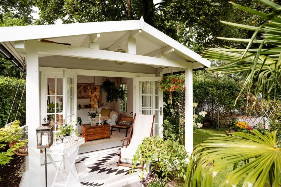 Gemütliche Schrebergarten Ideen: Ein weißes Gartenhaus mit offener Tür, Liegestuhl und Hängeampel lädt zum Verweilen ein.