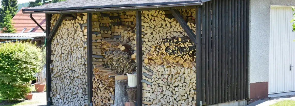 Ein Holzunterstand bauen: Gestapeltes Brennholz wartet unter einem Dach. Daneben eine Garage und grüne Büsche.