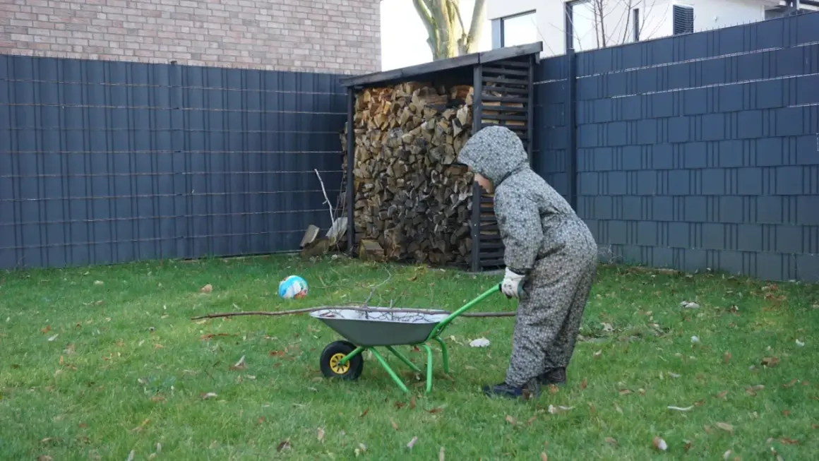 Kind schiebt Schubkarre mit Ästen am Zaun entlang. Gartengestaltung am Zaun entlang mit Holzstapel und Ball.