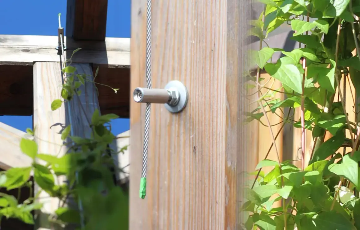 Selbstgebaute Rankhilfe aus Metall: Ein Drahtseil wird an einem Holzpfosten befestigt, um Kletterpflanzen Halt zu geben.