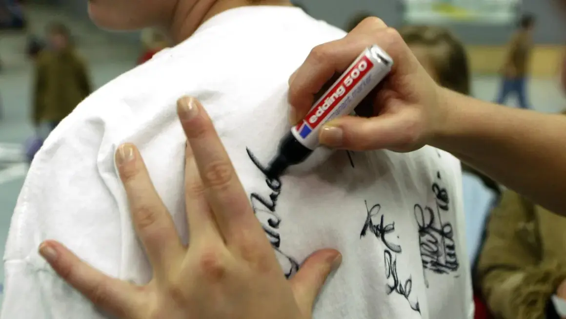 Eine Hand schreibt mit einem Edding-Filzstift Autogramme auf ein weißes T-Shirt. Tipps für hausmittel filzstift entfernen sind hier nicht zu sehen.