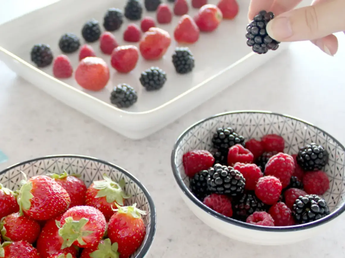 Frische Beeren, darunter Brombeeren, werden für das Einfrieren vorbereitet. Eine Hand hält eine Brombeere.